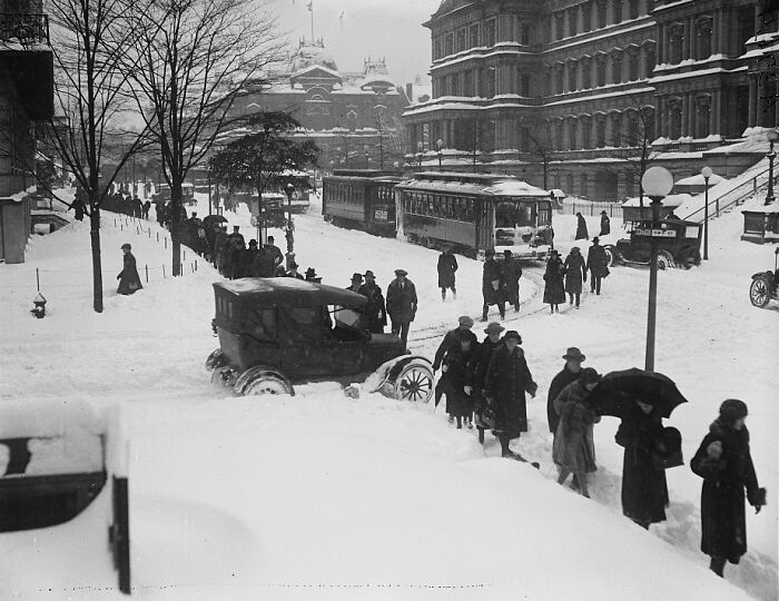 Snow-covered street in 1920s Washington, D.C. with pedestrians, vintage cars, and streetcars in a historic urban scene.