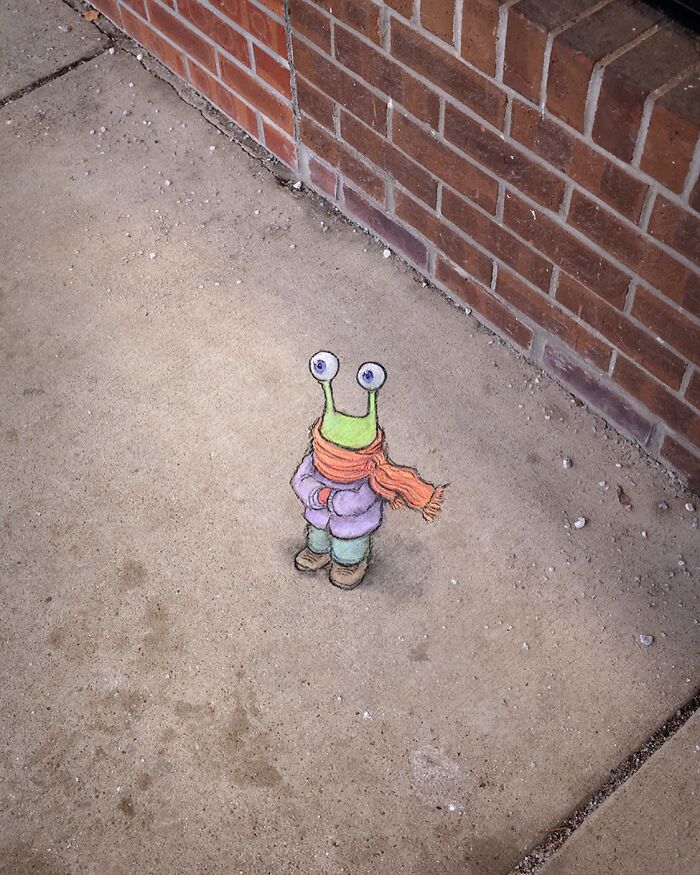Chalk art of a small green snail-like creature wearing a scarf and jacket on a sidewalk by a brick wall by David Zinn.