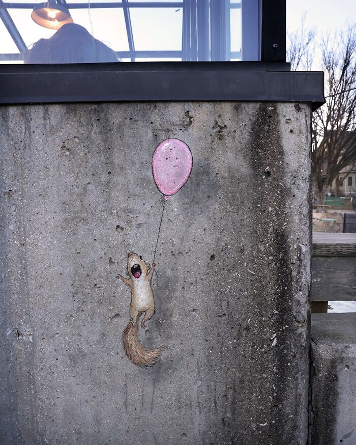 Chalk art by David Zinn showing a squirrel holding a pink balloon, blending with the concrete sidewalk wall.