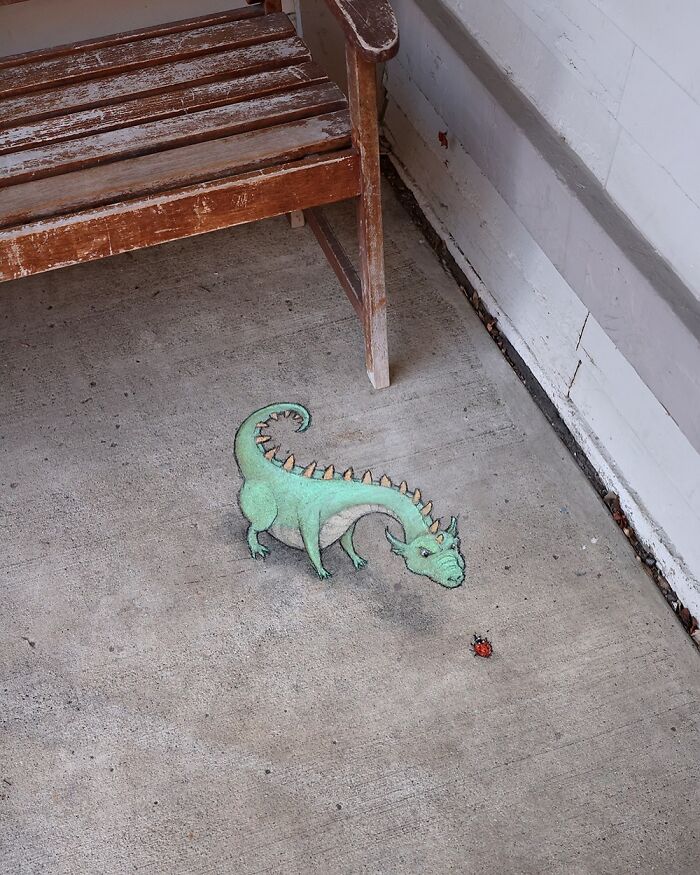 Chalk drawing by David Zinn of a small green dinosaur and a ladybug on a concrete sidewalk near a wooden bench.