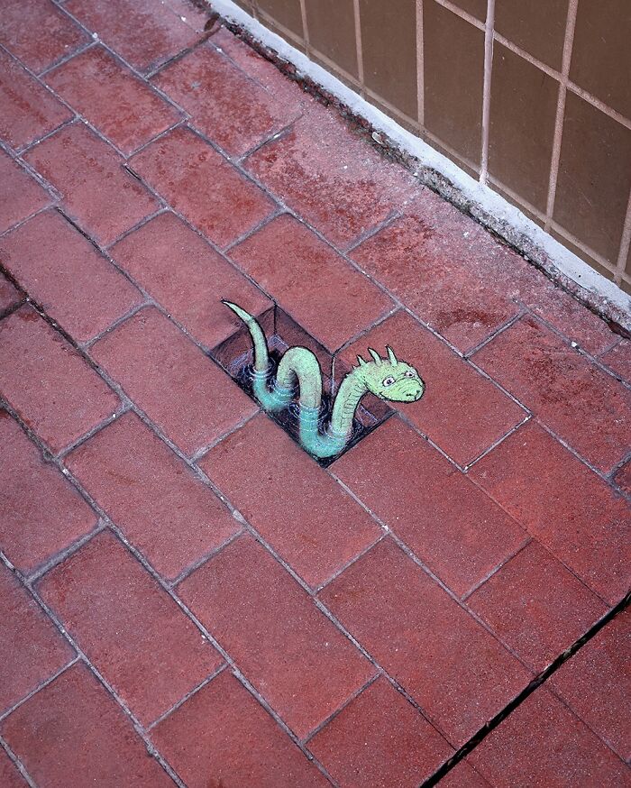 Chalk drawing by David Zinn of a whimsical green creature emerging from a gap in a red brick sidewalk.