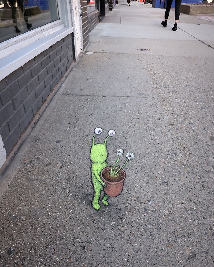 Chalk art by David Zinn showing a small green creature holding a pot with flowers on a city sidewalk.