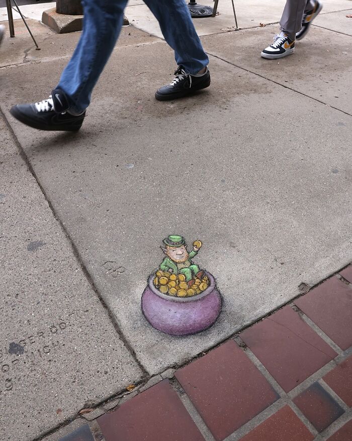 Sidewalk chalk art of a leprechaun in a pot of gold by David Zinn, with pedestrians walking nearby.