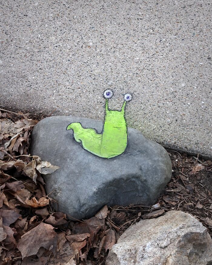 Chalk art by David Zinn featuring a whimsical green snail drawn on a rock near a concrete sidewalk and dry leaves.