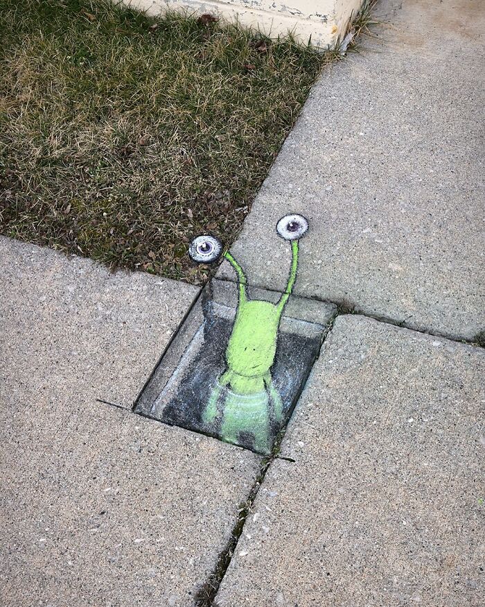 Chalk art by David Zinn showing a green snail with large eyes emerging from a sidewalk crack near grass and a building corner.