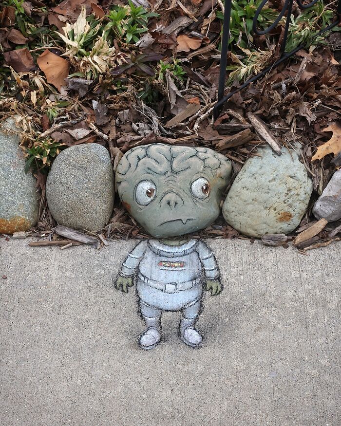 Chalk art by David Zinn of a small alien character with a rock as a head on a sidewalk surrounded by leaves and stones.