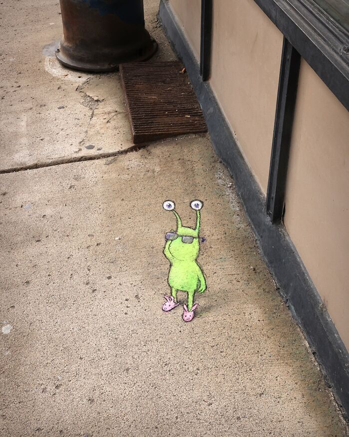 Chalk art by David Zinn showing a green creature with glasses and pink slippers on a sidewalk near a building.