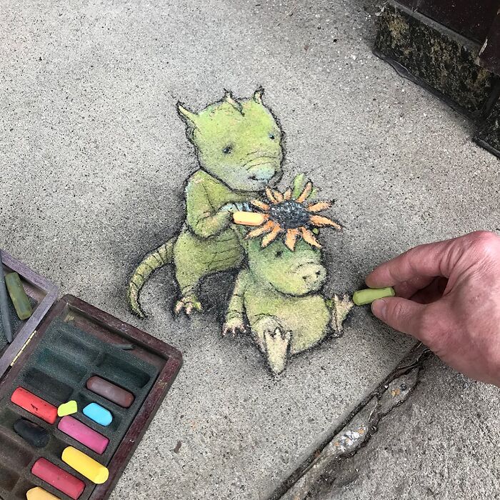 Chalk art by David Zinn showing two green creatures on a sidewalk, one with a flower on its head, being drawn by hand.