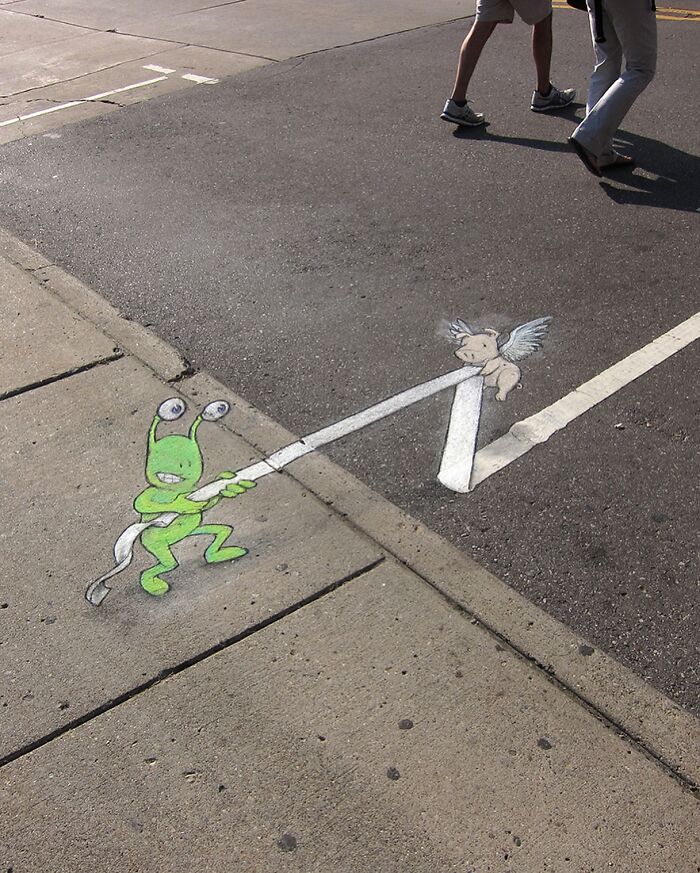 Sidewalk chalk art by David Zinn showing a green alien pulling a winged pig with a rope on the street.