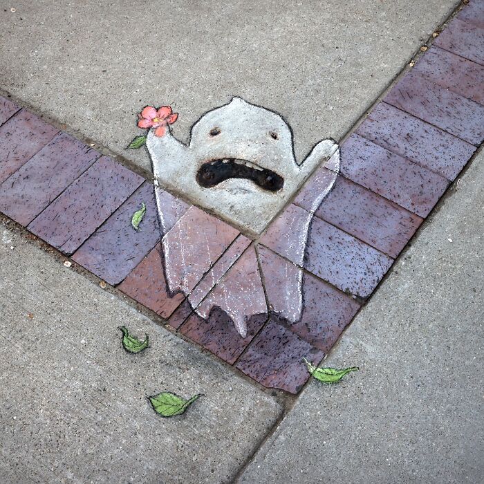 Chalk art by David Zinn of a small ghost holding a pink flower on a sidewalk corner with scattered green leaves.