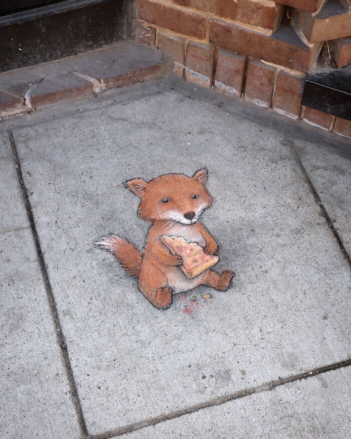 Chalk art by David Zinn showing a cute fox holding a slice of pizza drawn on a concrete sidewalk corner.