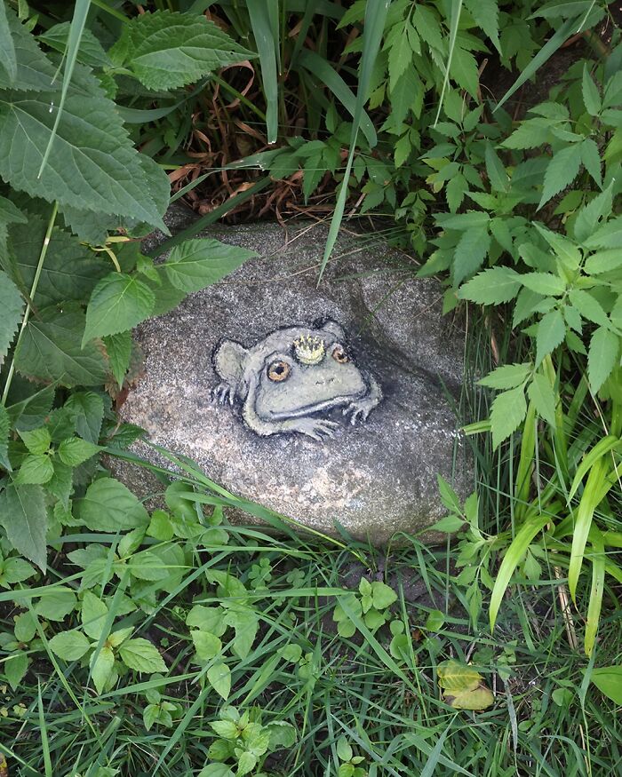 Chalk art by David Zinn depicting a frog with a small crown on a rock surrounded by green plants and grass.