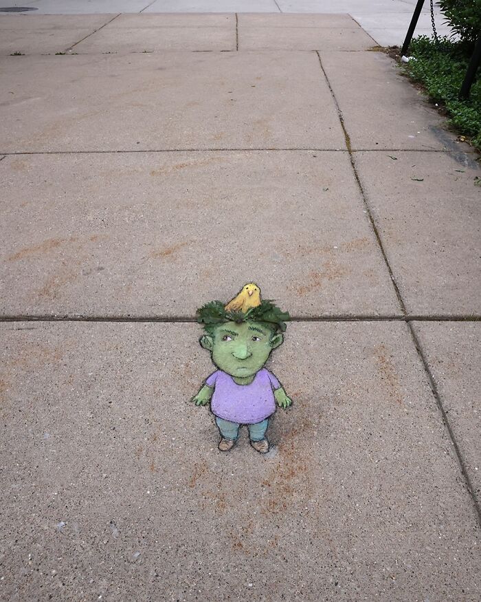 Chalk drawing by David Zinn on sidewalk of a small green character with a bird on its head surrounded by leaves.