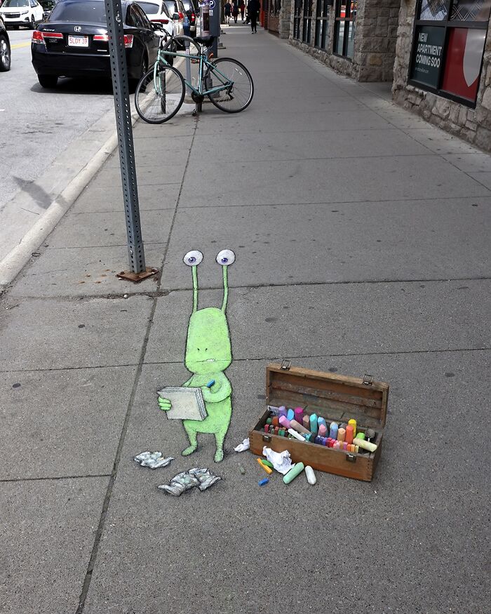 Chalk art by David Zinn features a green two-eyed creature sketching beside a box of colorful sidewalk chalk.