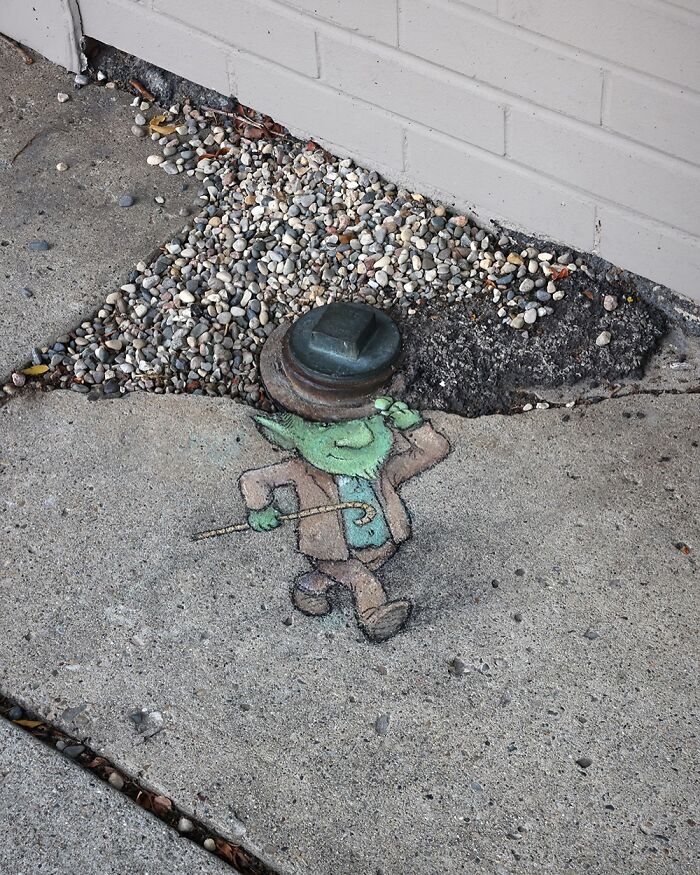 Chalk drawing by David Zinn on sidewalk depicting a small green character tipping a hat near gravel and concrete.
