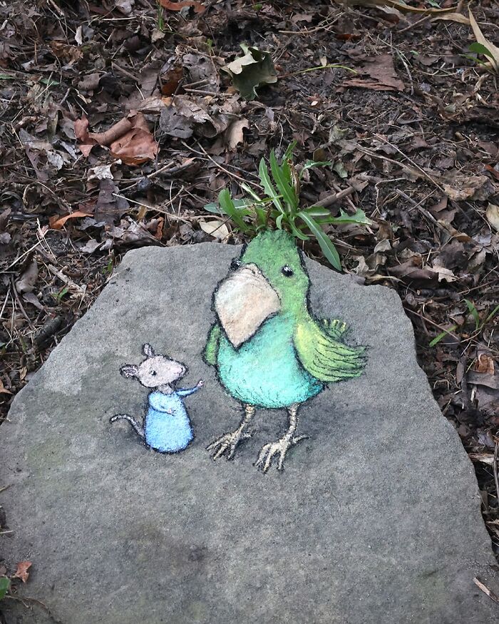 Chalk art by David Zinn of a mouse and a green bird on a stone surrounded by leaves and plants outdoors.