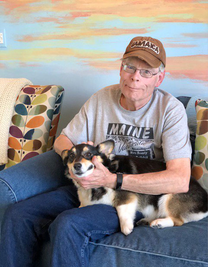 Stephen King sitting on sofa with dog, wearing a Maine t-shirt and Jamaica cap amid Epstein List conspiracy theories. - 1