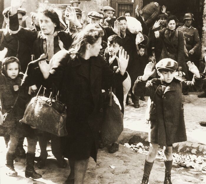 Group of civilians, including women and children, with raised hands under guard in a somber historical moment.
