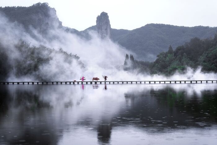 Misty mountain landscape in Asia with two people and a cow crossing a narrow bridge, capturing the soul of Asia in a stunning photo.