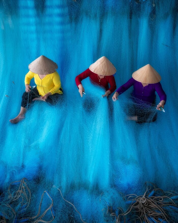 Three Asian women wearing traditional hats are mending vibrant blue fishing nets, capturing the soul of Asia in a stunning photo.