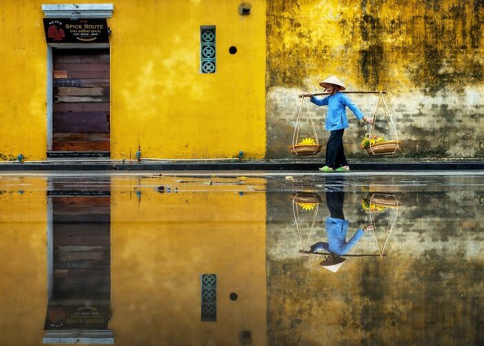 Person wearing a traditional hat carrying baskets walking past a yellow wall with a reflection on wet ground, capturing the soul of Asia.