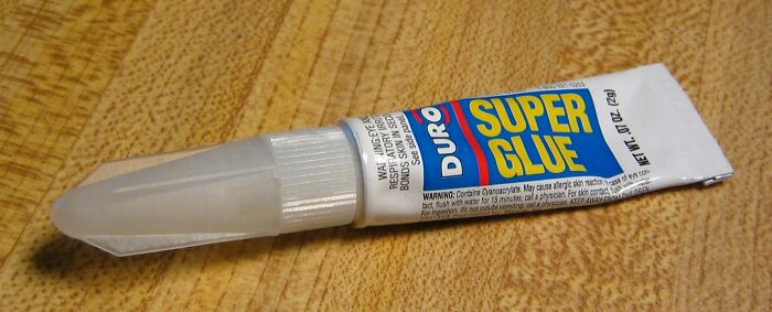 Tube of super glue with an attached plastic applicator cap placed on a wooden surface for ingenious inventions.