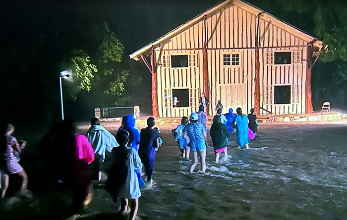 People wade through floodwaters at night near a wooden building during Texas floods affecting family of Travis Kelce's boss.