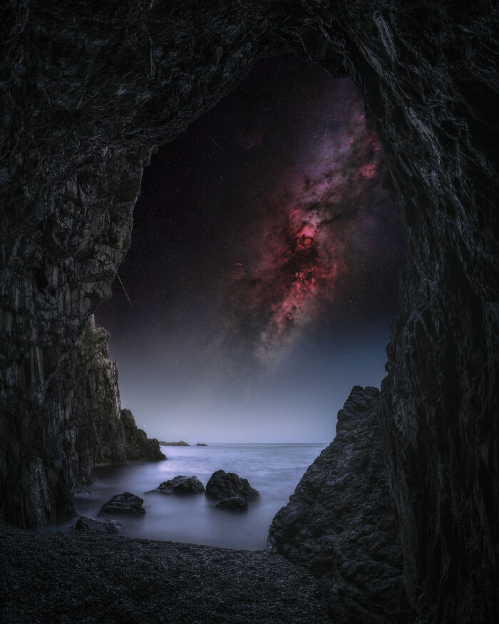 View of the Milky Way galaxy over calm sea rocks framed by a dark cave entrance in breathtaking space photography.
