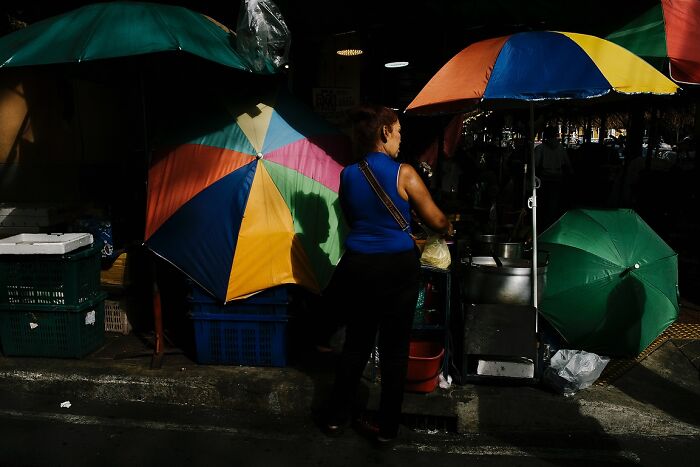 Woman standing under colorful umbrellas preparing food, captured in powerful street photos of India by photographer Rohit Vohra.