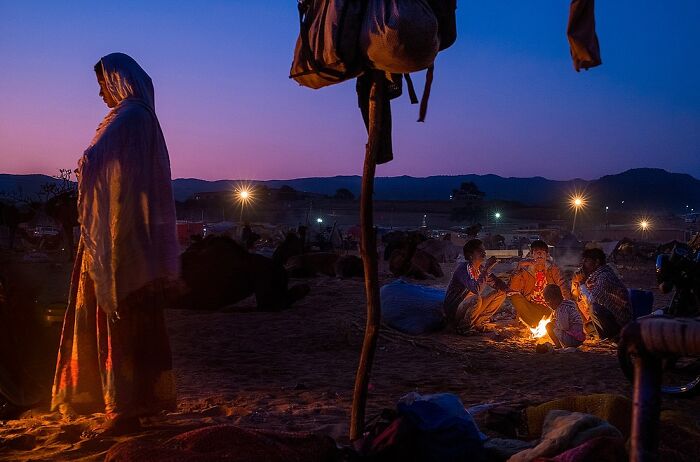 Woman in traditional attire stands near a group by fire at dusk in a powerful street photo capturing the soul of India.