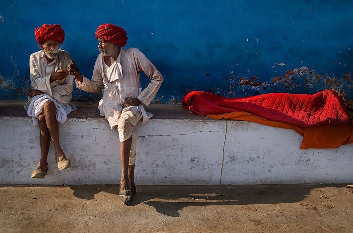 Two Indian men wearing traditional clothing and red turbans seated against a blue wall in a powerful street photo capturing the soul of India.