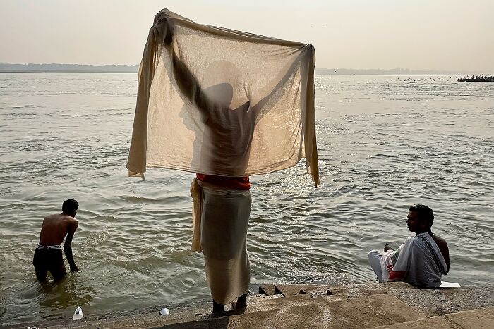 Man holding translucent cloth by riverbank with two men bathing, a powerful street photo capturing the soul of India.