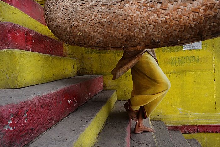 Woman carrying a large basket walking barefoot up colorful stairs in a powerful street photo capturing the soul of India.