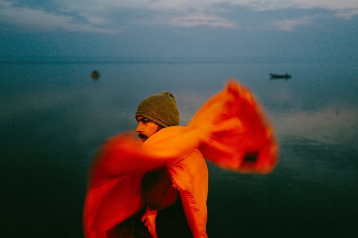 Man wrapped in a bright orange shawl near calm water, one of the powerful street photos capturing the soul of India.