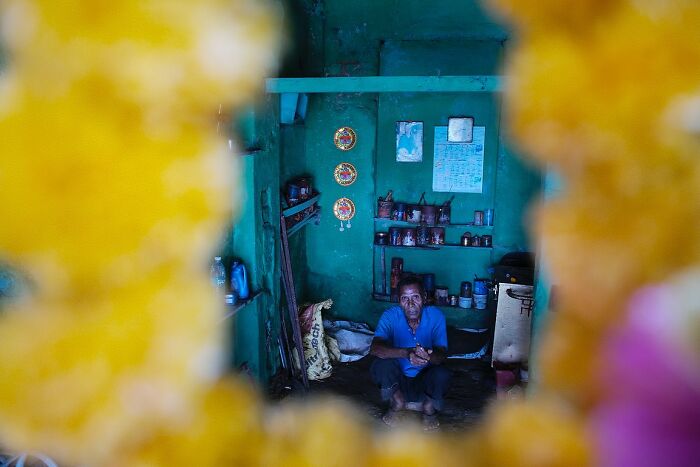 Elderly man sitting inside a rustic room, framed by vibrant yellow garlands in powerful street photos capturing the soul of India.