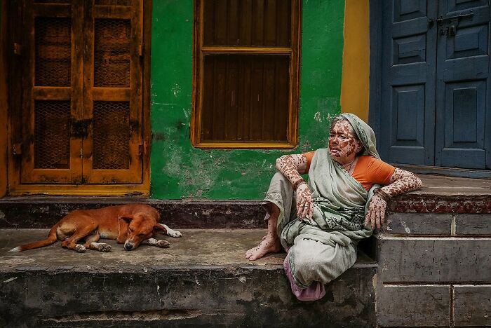 Elderly woman with henna tattoos sitting by colorful doors with a sleeping dog in a powerful street photo of India