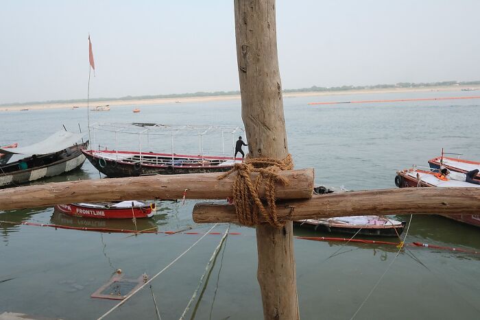 Wooden pole and ropes tied by the riverbank with boats floating on water in a powerful street photo of India.