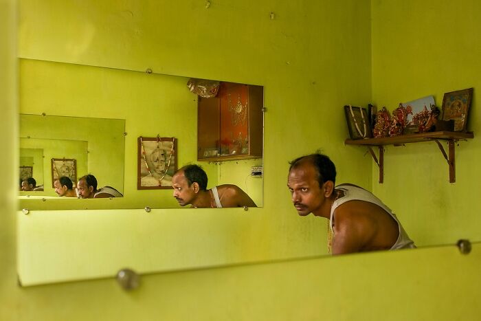 Man leaning forward inside a yellow room with multiple reflections in mirrors, a powerful street photo capturing the soul of India.