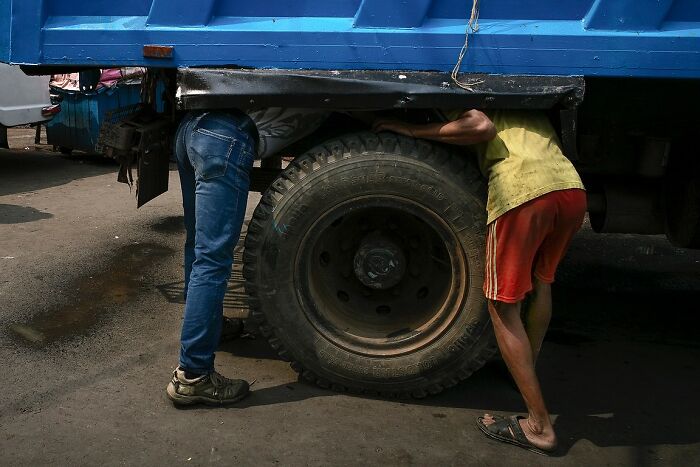 Two men working under a truck tire on a street, captured in powerful street photos of India by Rohit Vohra.