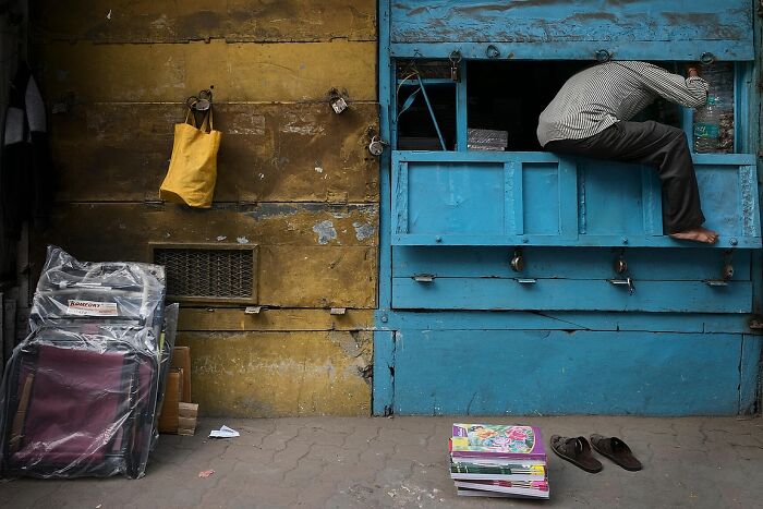 Man climbing into a blue street stall against a yellow wall, captured in powerful street photos of India.