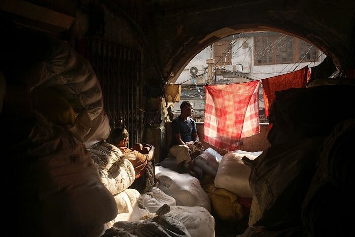 Two men sitting among sacks in a dimly lit space with red cloth hanging, powerful street photos from India perspective.