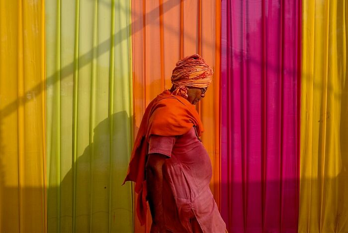 Man in orange and pink attire walking past vibrant striped yellow, orange, and pink wall in powerful street photos of India.