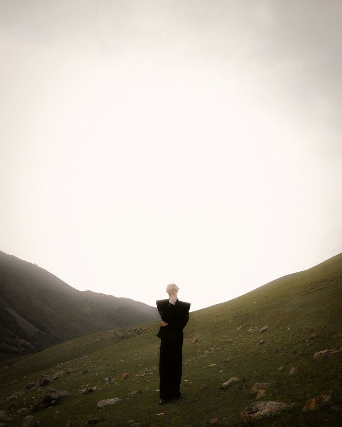 Person dressed in black standing thoughtfully on a green hillside, showcasing breathtaking color photography.