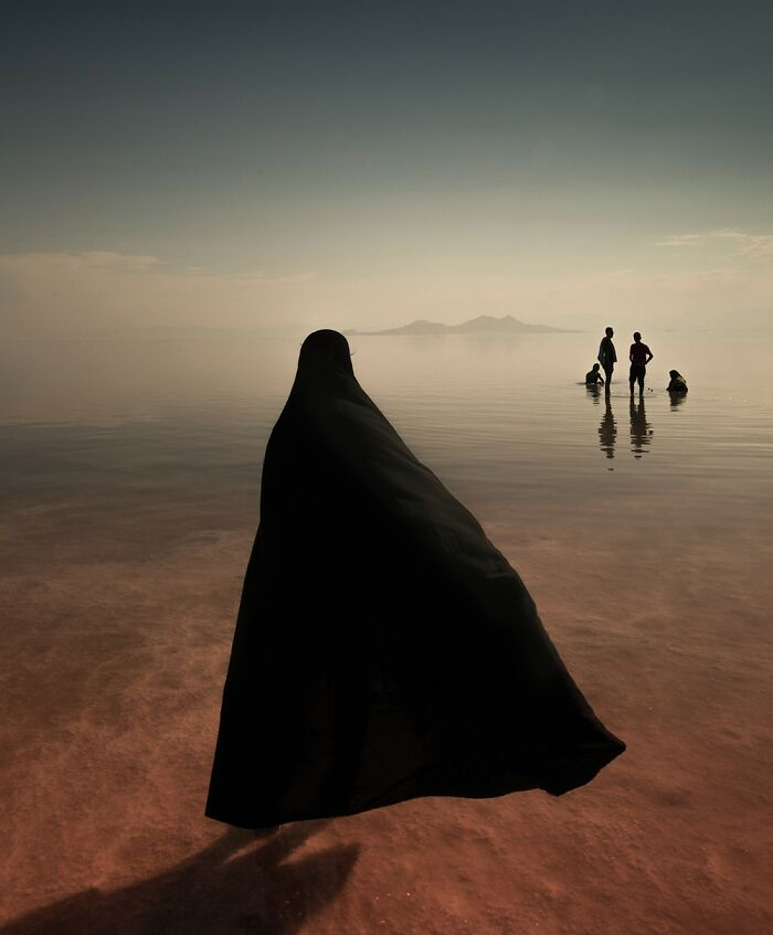 A person in black cloak standing in shallow water with several people in distance, showcasing breathtaking color photography.