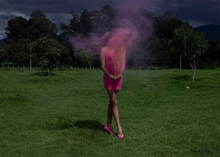 Woman in pink dress and heels standing in green field with pink powder clouding her face, showcasing stunning color photography.