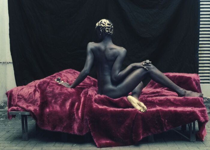 Artistic color photography of a n**e figure with painted head, posed on a red textured fabric, showcasing creative composition.