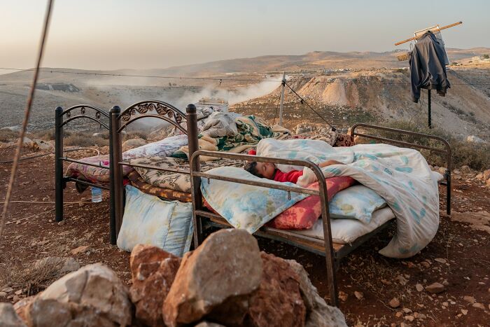 Child sleeping wrapped in blankets on an outdoor bed amidst a rugged landscape captured in color photography award winner.