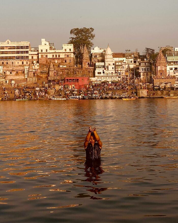 Person performing a ritual in the river with historic Asian city buildings densely packed along the shore in the background.