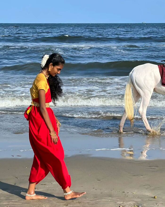 Young woman in traditional Asian dress walking barefoot on a beach near ocean waves with a white horse in the background.