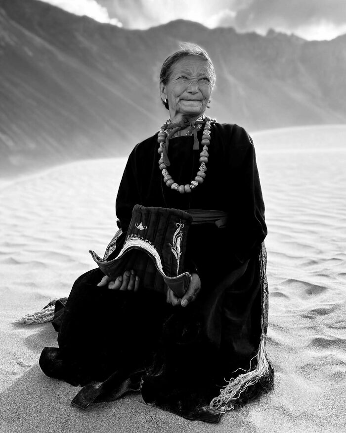 Elderly Asian woman in traditional attire sitting on sand with mountains in the background, showcasing Asia photography.